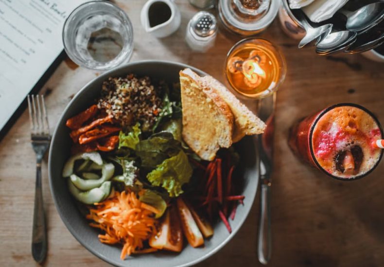 Top view of healthy salad with assorted vegetables and toasted bread placed on wooden table with exotic fresh drink and cutlery